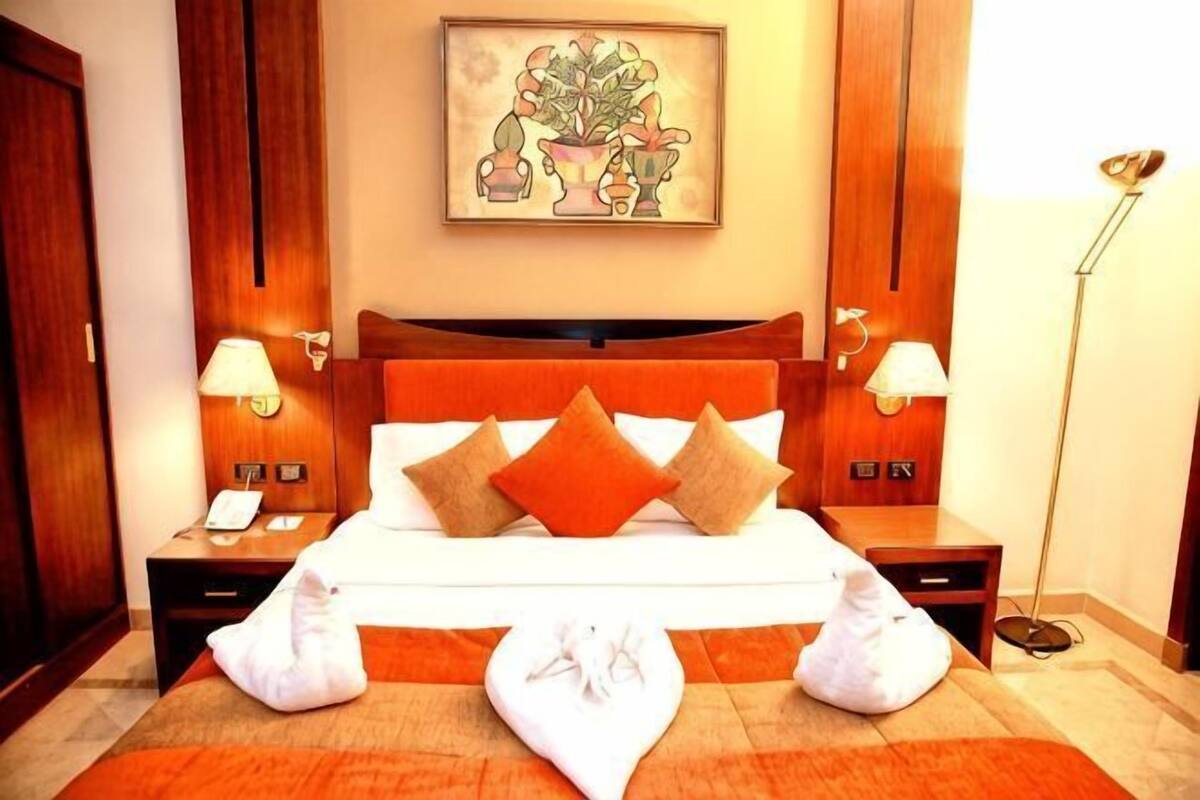 A hotel room with a bed, two bedside tables, a nightstand, a lamp, a painting, and a floor lamp.