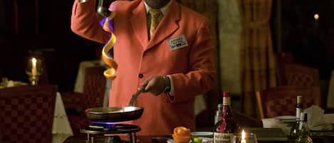 A man in a red suit cooking with a traditional stove, surrounded by dining tables set with plates, glasses, and candles.