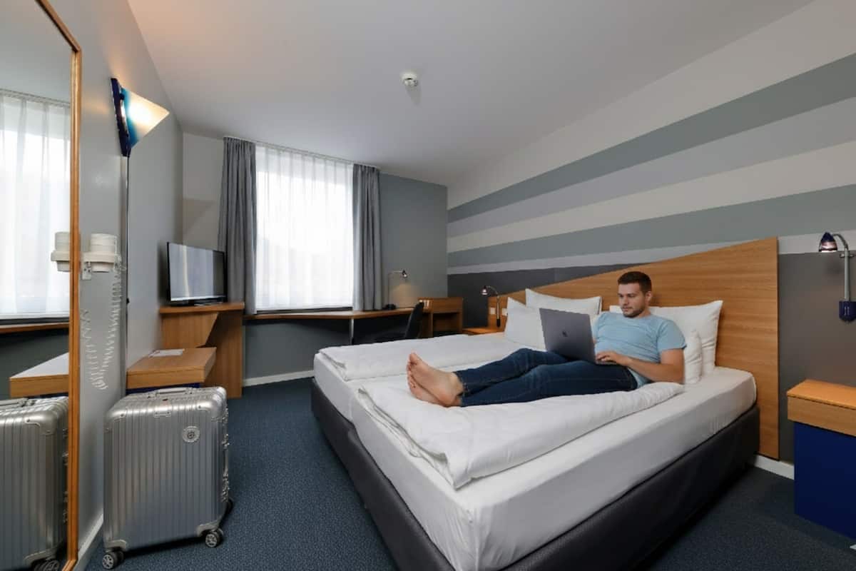 Habitació d'hotel amb un llit, un escriptori, una cadira, un televisor, una maleta i una làmpada.