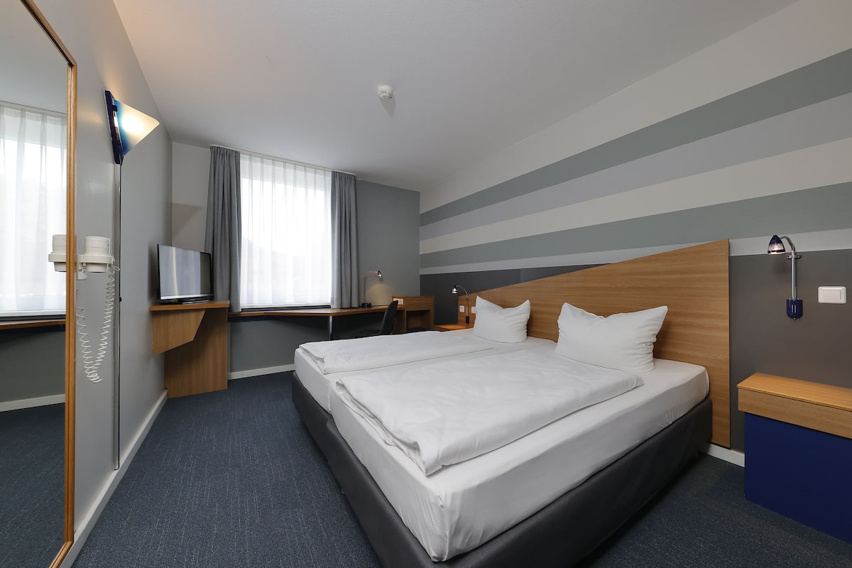 A hotel room with a large bed, a desk with a chair, a TV, and a window with curtains.