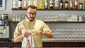 Uma pessoa vestindo uniforme de chef limpando um copo com um pano e uma garrafa em um bar, com prateleiras cheias de garrafas de bebidas e copos.