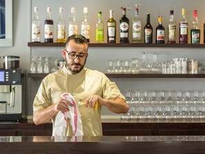 A person in a chef's uniform cleaning a glass with a cloth and a bottle in a bar setting with shelves of liquor bottles and glasses.