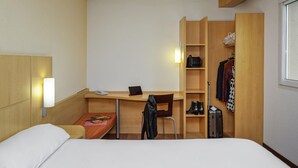 Quarto de hotel compacto com cama, escrivaninha com cadeira, pequena mesa com telefone e tablet, guarda-roupa com roupas penduradas e sapateira.
