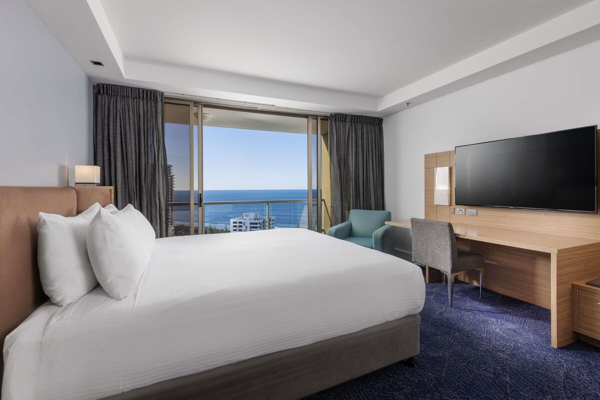 A hotel room with a large bed, a desk with a flat-screen TV, a chair, and a view of the ocean.