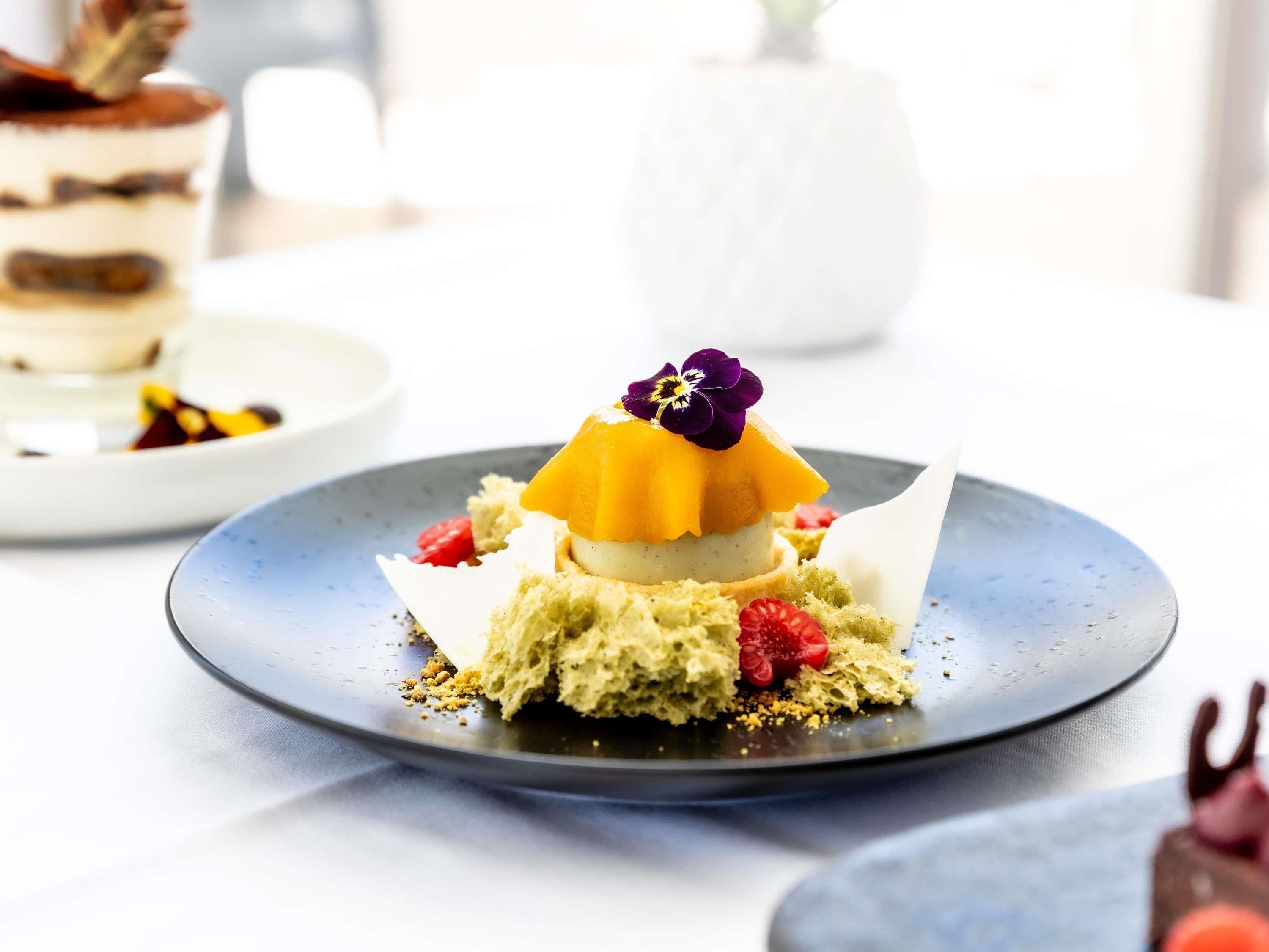 A dessert on a plate with a layered cake, a yellow dome, and a purple flower on top.