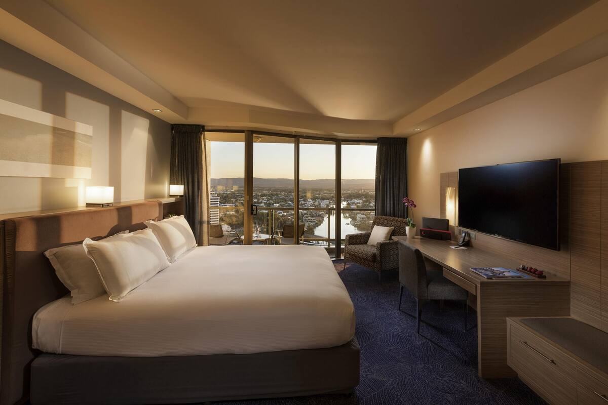 A hotel room with a large bed, a desk with a flat-screen TV, a chair, and a view of the city through large windows.
