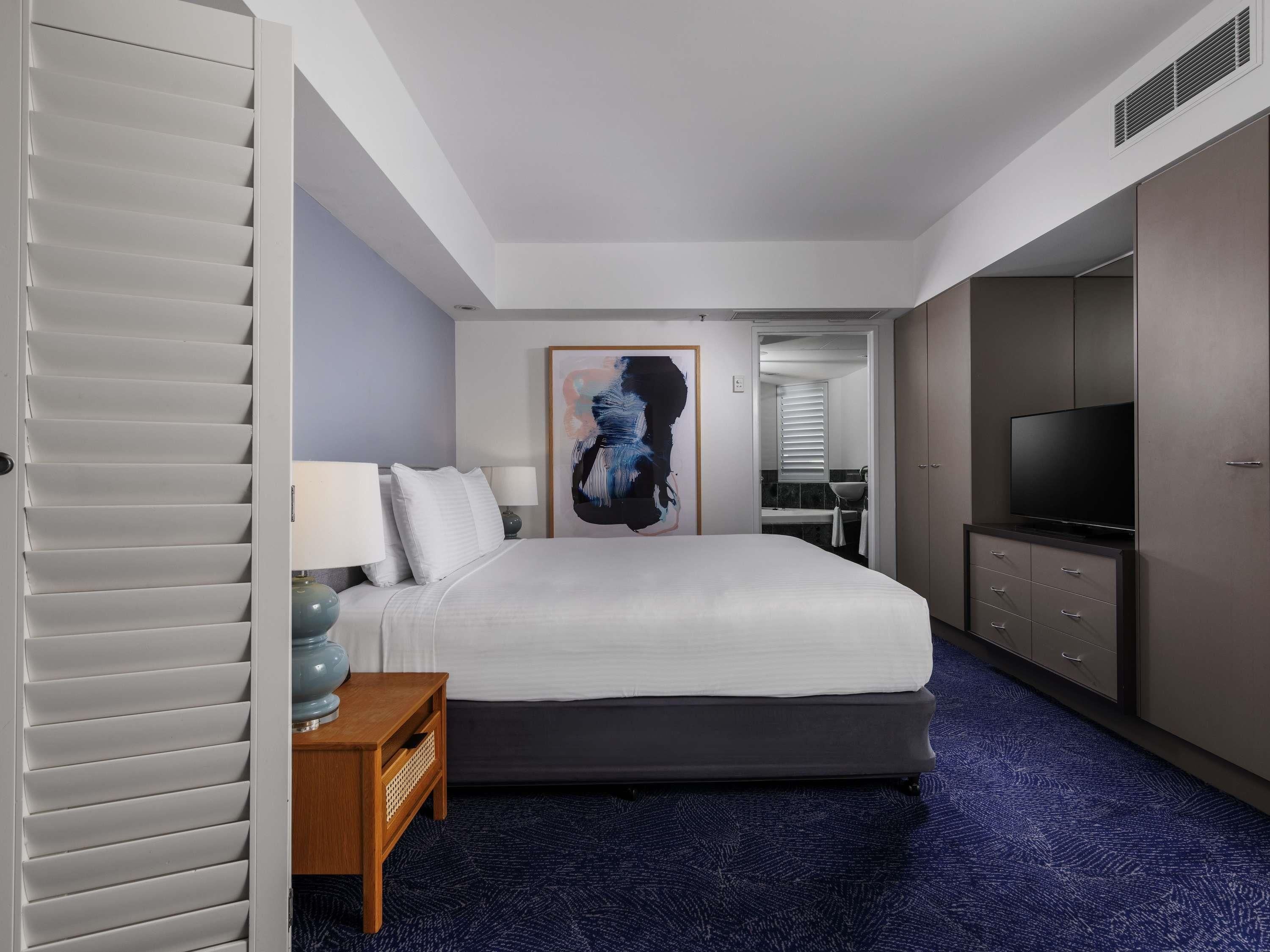 Habitación de hotel con cama, mesita de noche, lámpara, obra de arte, televisor, armario y baño a la vista a través de una puerta abierta.