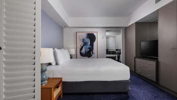 Habitación de hotel con cama, mesita de noche, lámpara, obra de arte, televisor, armario y baño a la vista a través de una puerta abierta.