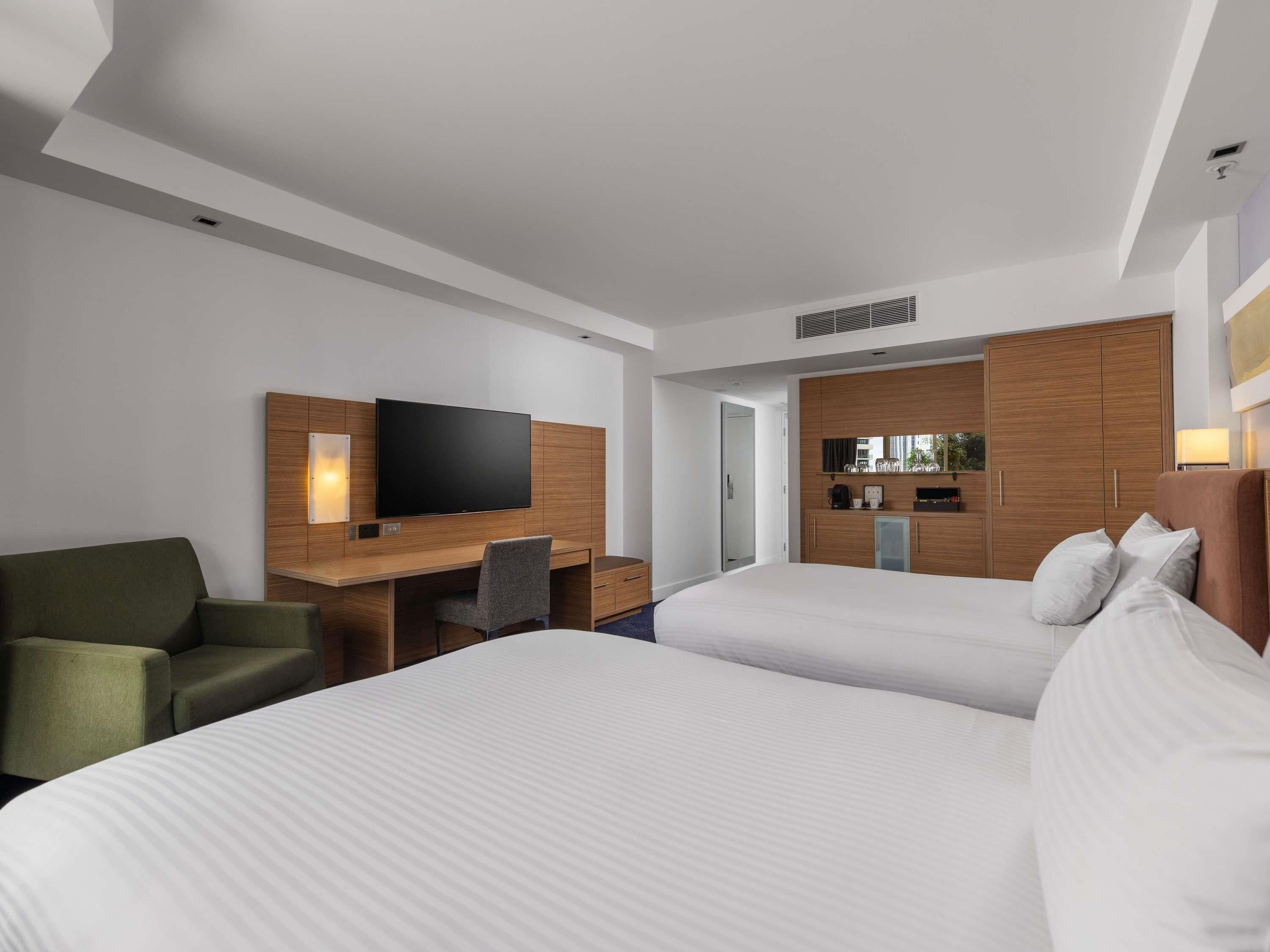 A hotel room with two beds, a desk with a flat-screen TV, a green armchair, and a minibar.