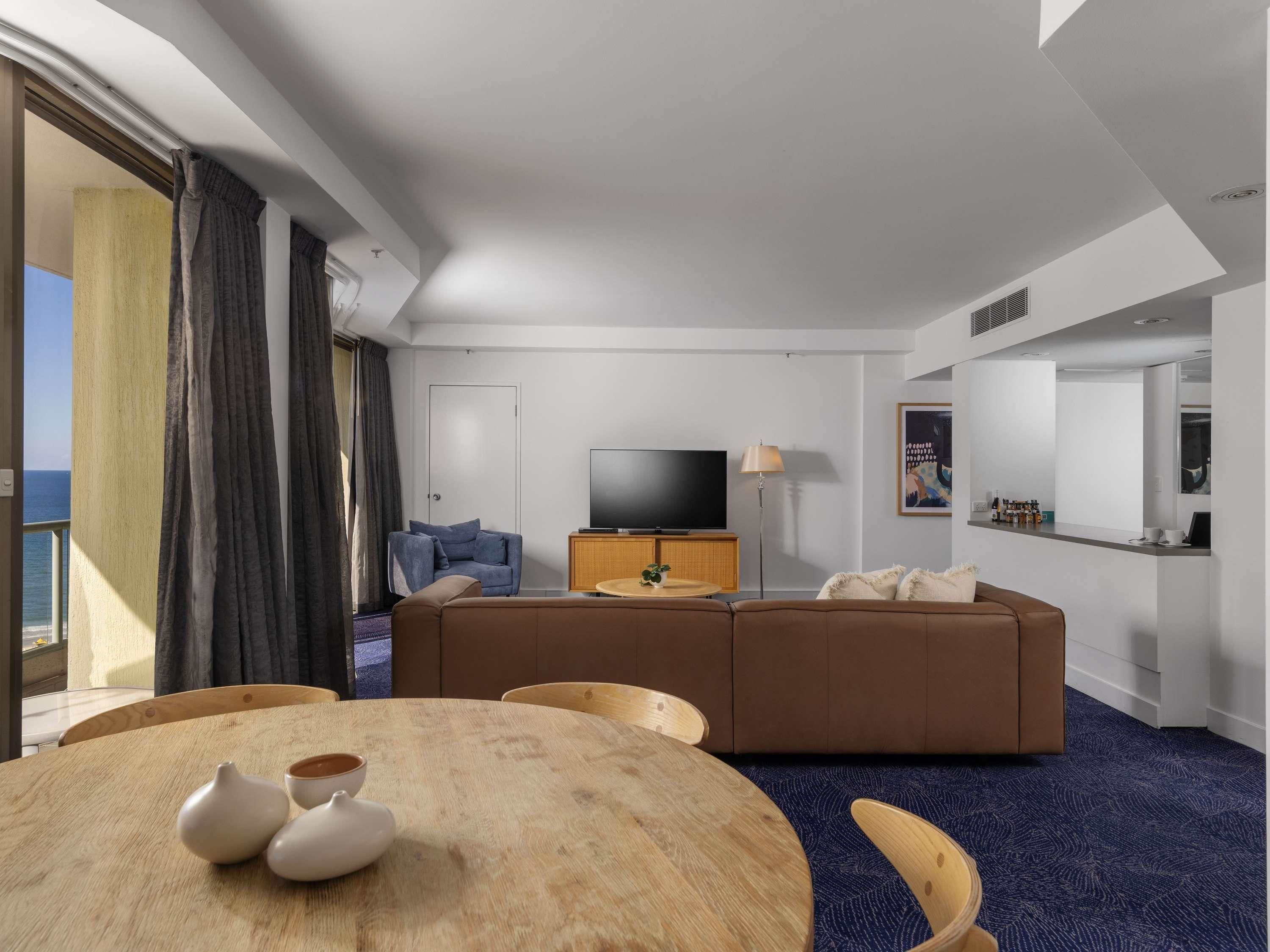 A modern hotel room with a large window, a flat-screen TV, a sofa, a dining table with chairs, and a minibar.