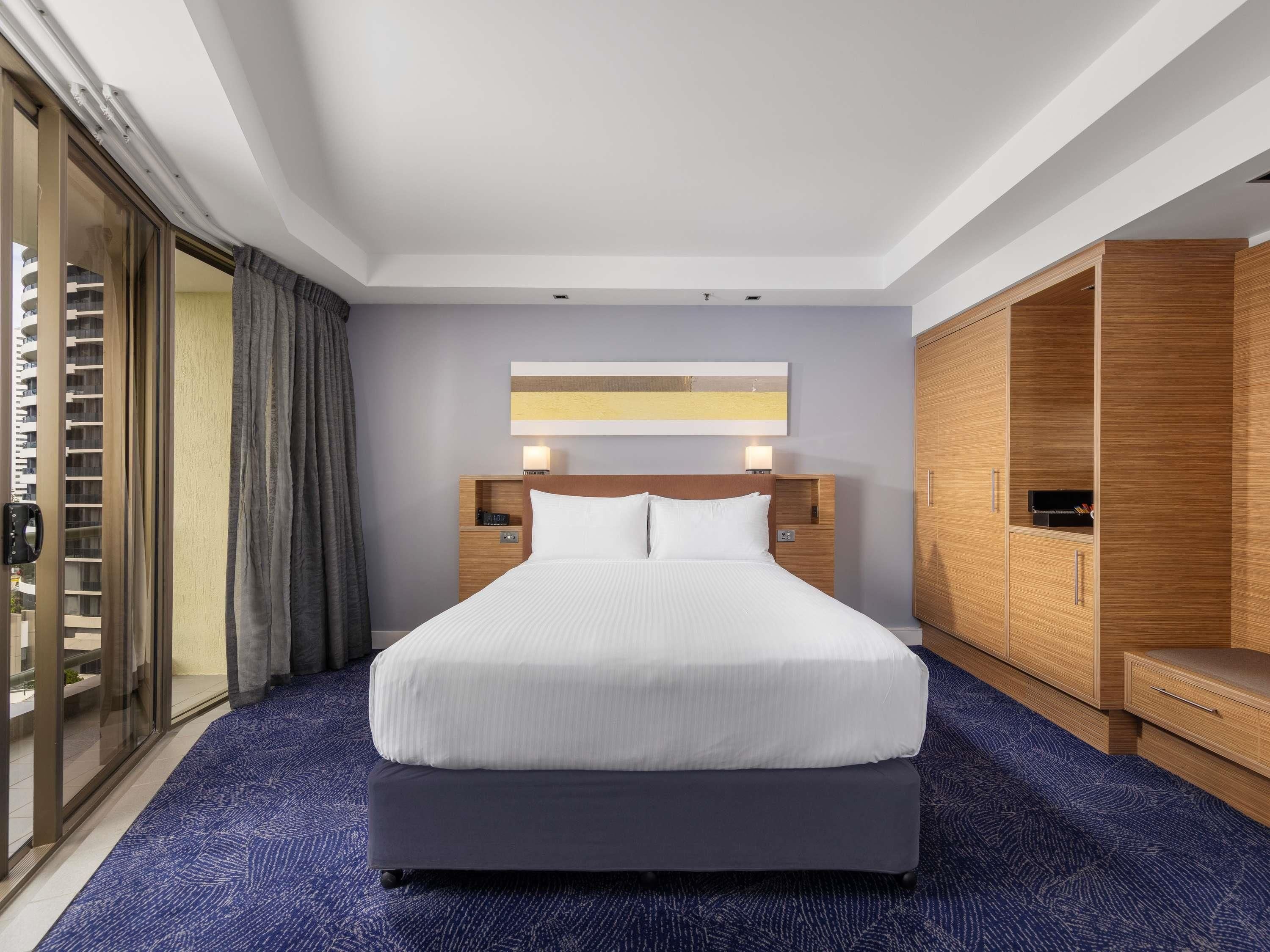 A hotel room with a large bed, a wooden wardrobe, a nightstand, and a view through a glass door.