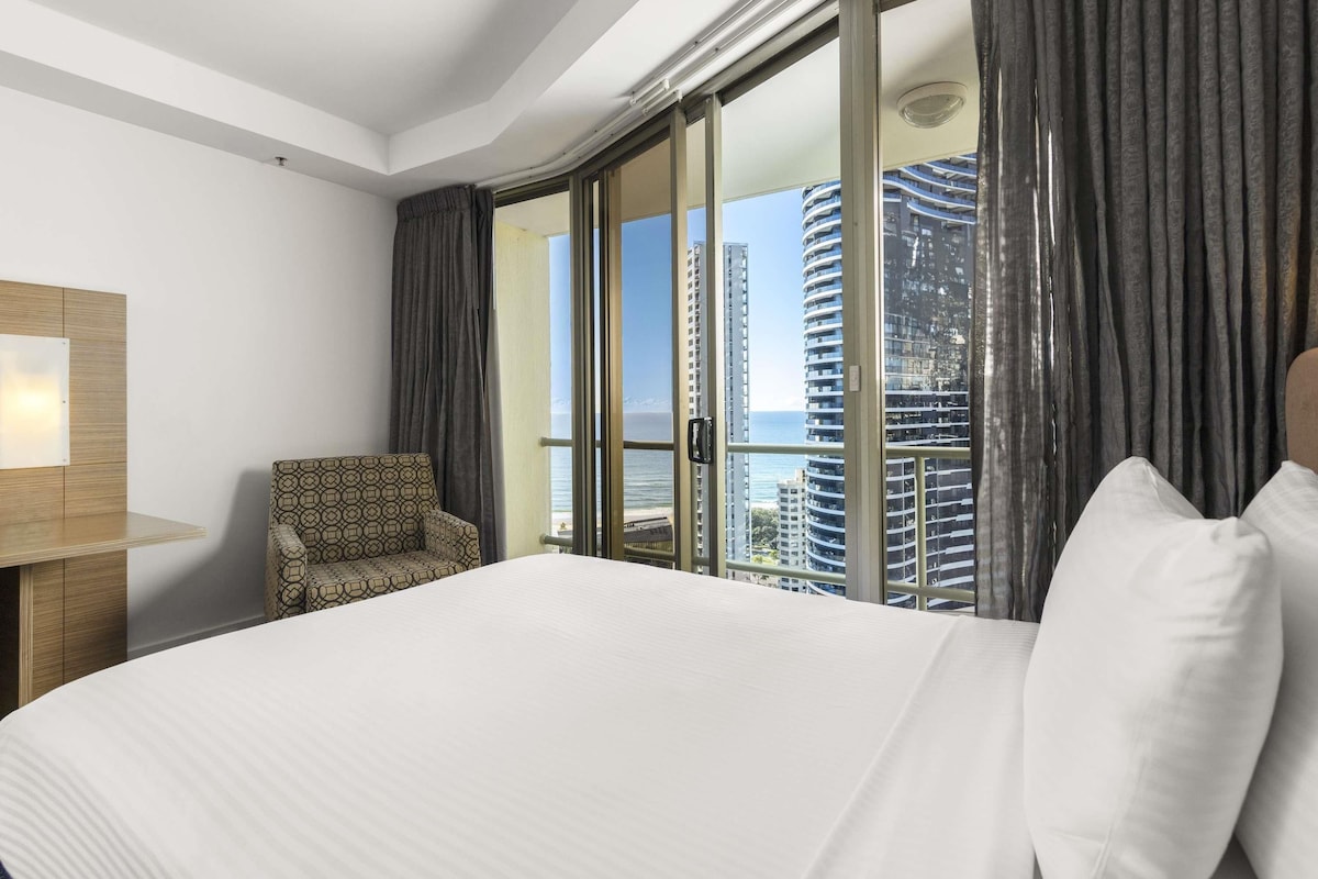 A hotel room with a large bed, a chair, and a view of tall buildings and the sea.