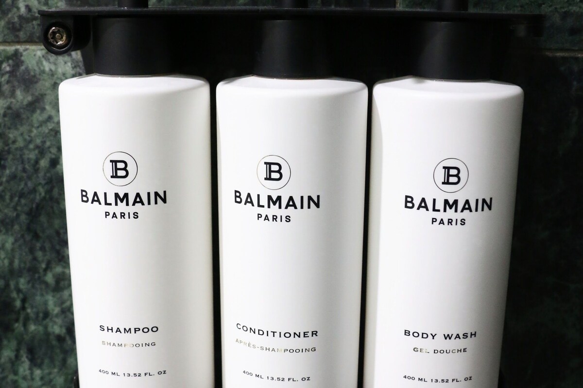 Trois produits Balmain Paris : un shampooing, un après-shampooing et un gel douche, présentés chacun dans un flacon blanc doté d’un distributeur noir, le tout fixé sur un support noir adossé à un mur en marbre.