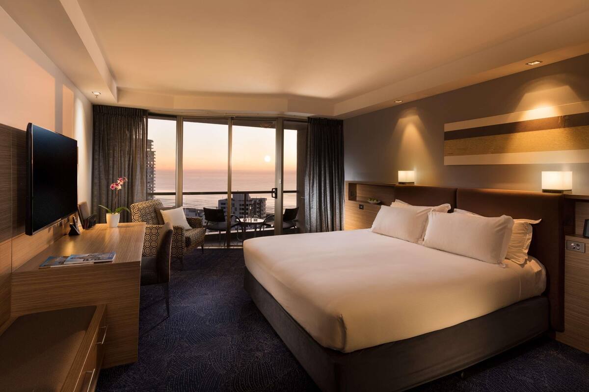 Une chambre d’hôtel dotée d’un grand lit, d’un bureau avec un téléviseur à écran plat, d’un coin salon avec une table et des chaises, et offrant une vue sur le coucher de soleil sur l’océan.