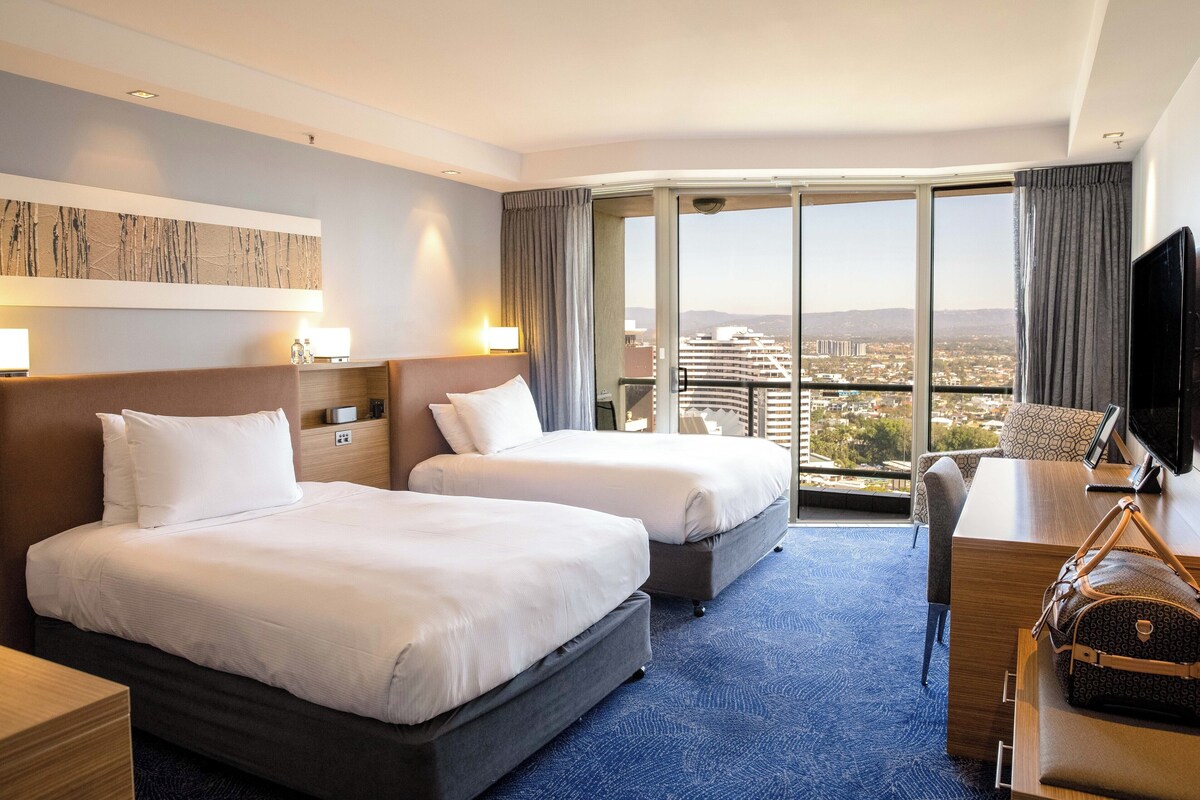 Une chambre d’hôtel avec deux lits, un bureau, une télévision et une grande fenêtre donnant sur la ville.
