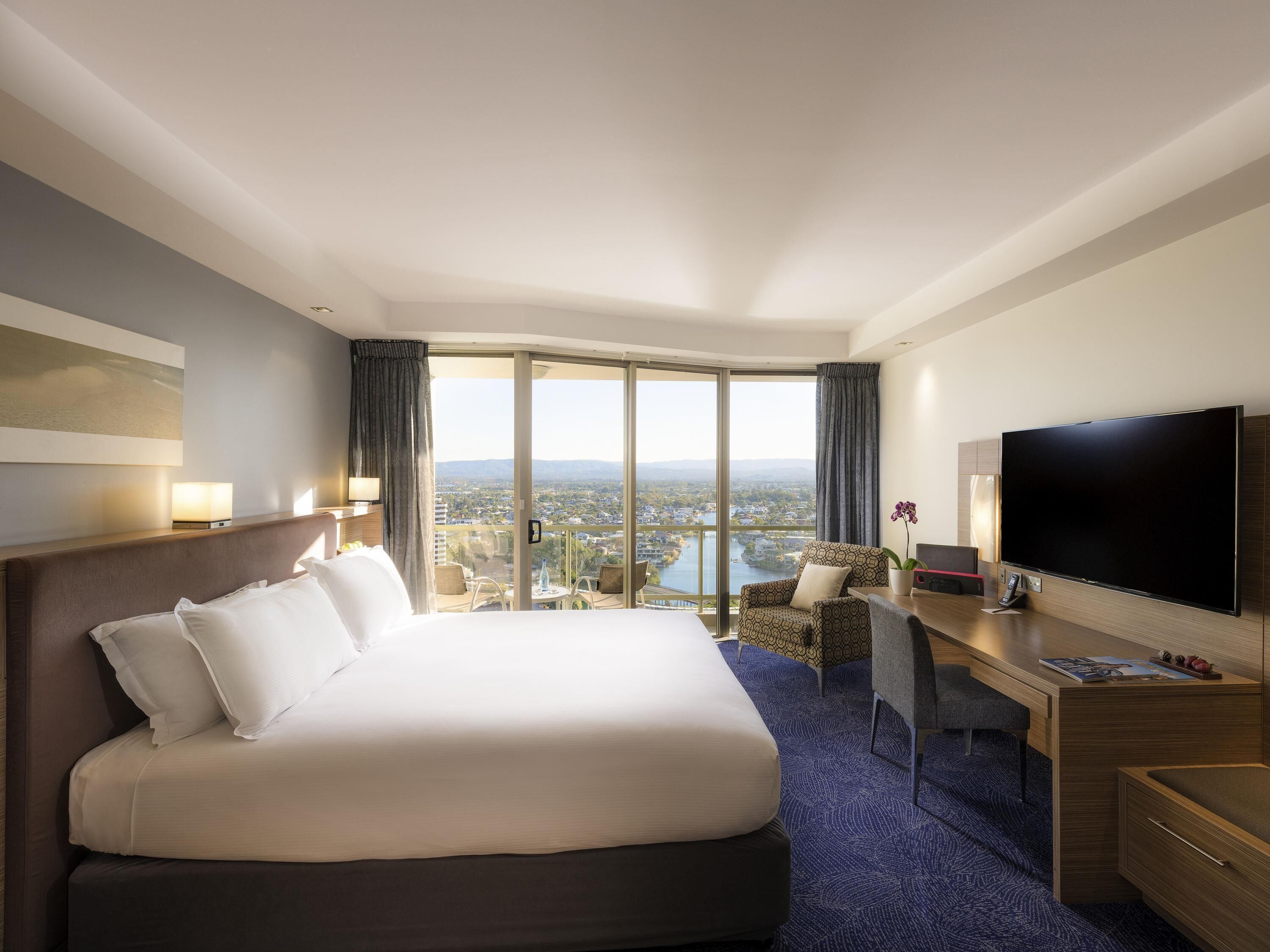 A hotel room with a large bed, a desk with a flat-screen TV, a chair, and a view of a cityscape through large windows.