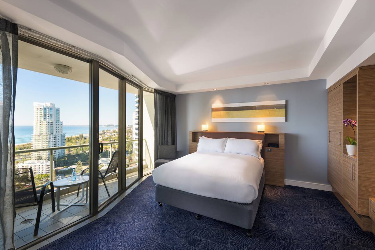 Une chambre d’hôtel avec un grand lit, une vue sur la ville, un balcon avec une table et des chaises, et une armoire en bois.