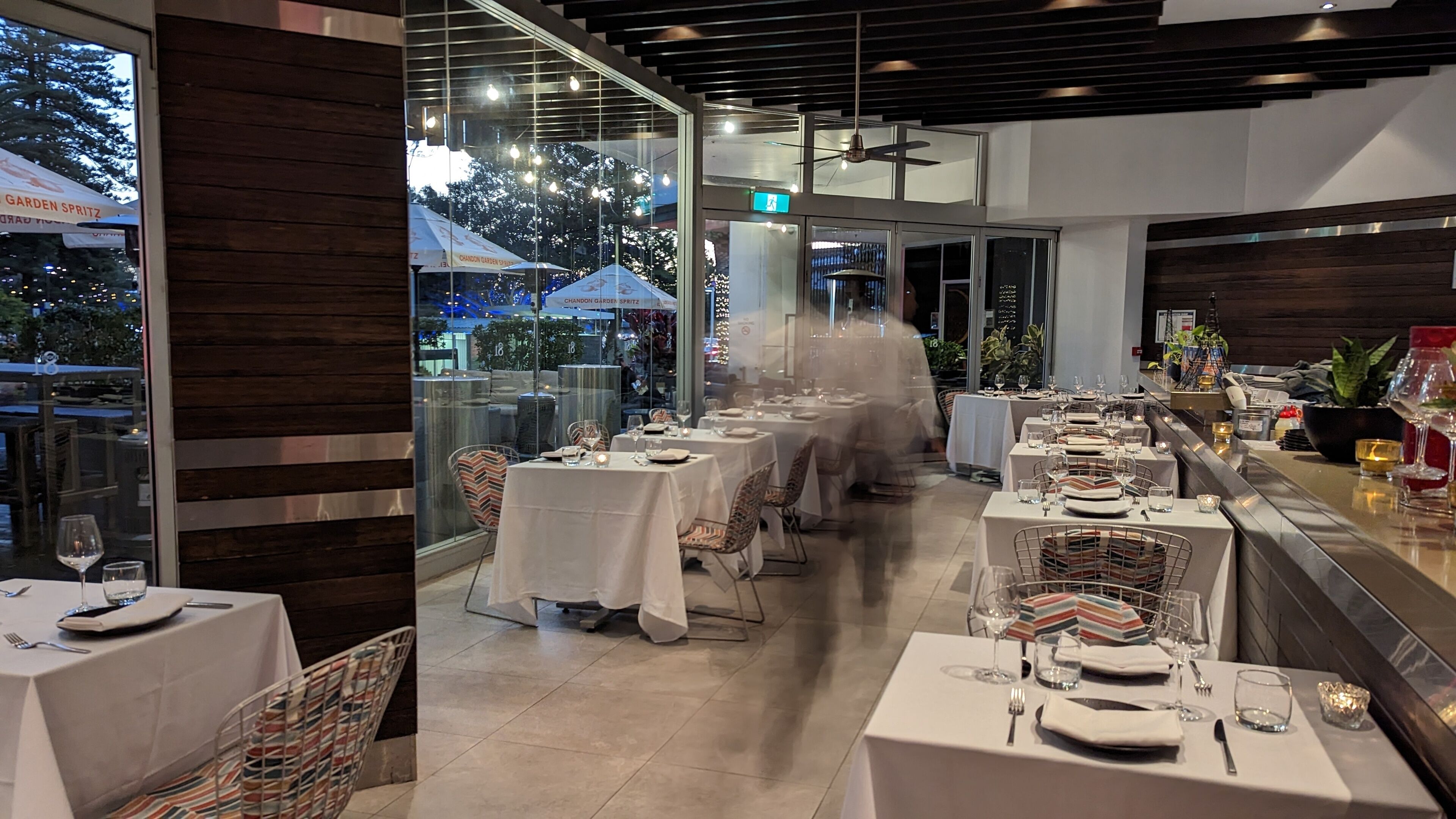 Un restaurante con varias mesas dispuestas para comer, una zona de bar con mostrador, y vistas del área exterior a través de grandes ventanales.