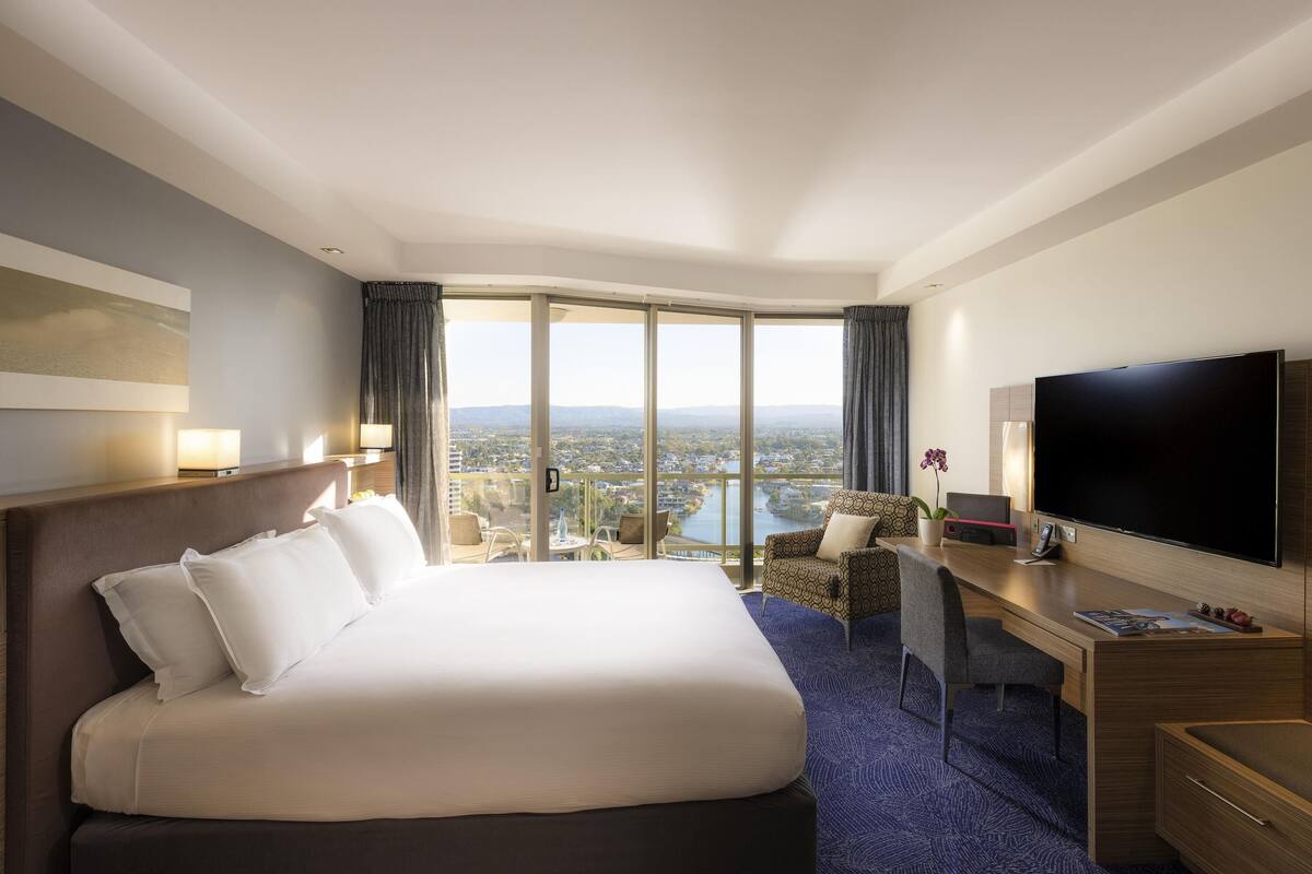 A hotel room with a large bed, a desk with a flat-screen TV, a view of a cityscape through large windows, and a balcony with a table and chairs.