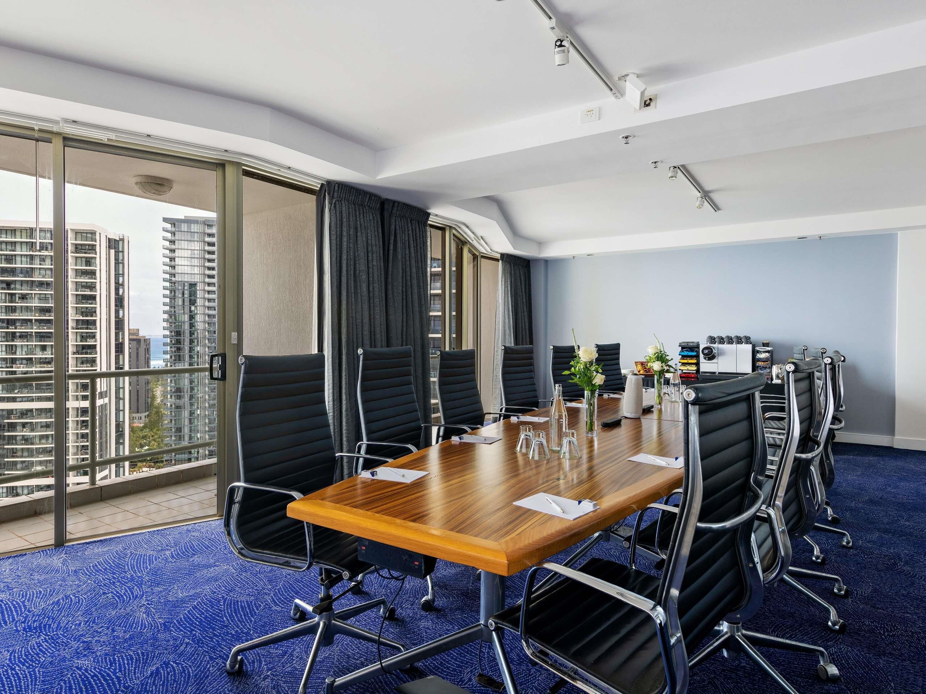 A conference room with a wooden table, black chairs, a blue carpet, and a city view through large windows.