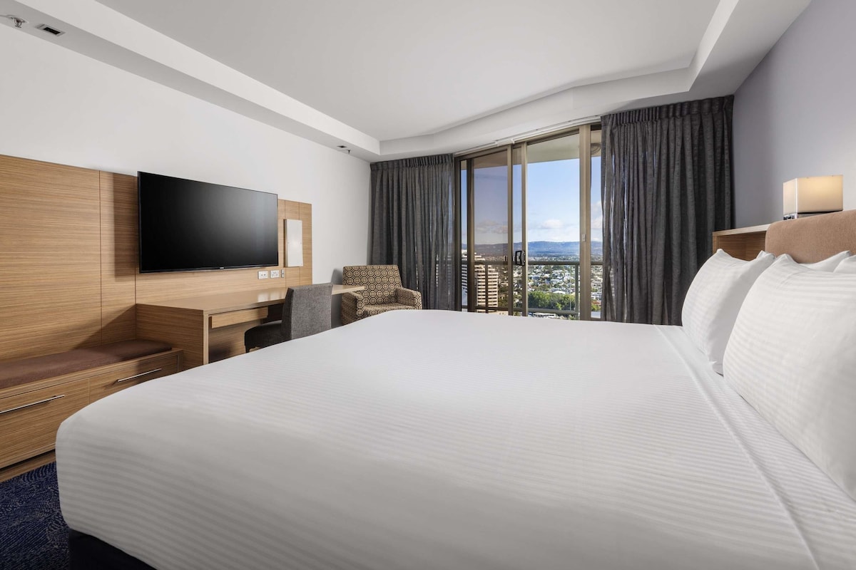 A hotel room with a large bed, a flat-screen TV mounted on the wall, a desk with a chair, and a view of the city through a large window.