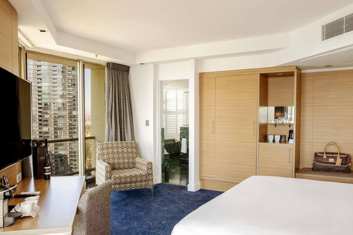 Une chambre d’hôtel avec un lit, un bureau avec une télévision, une chaise, une armoire et une vue sur des immeubles de grande hauteur.