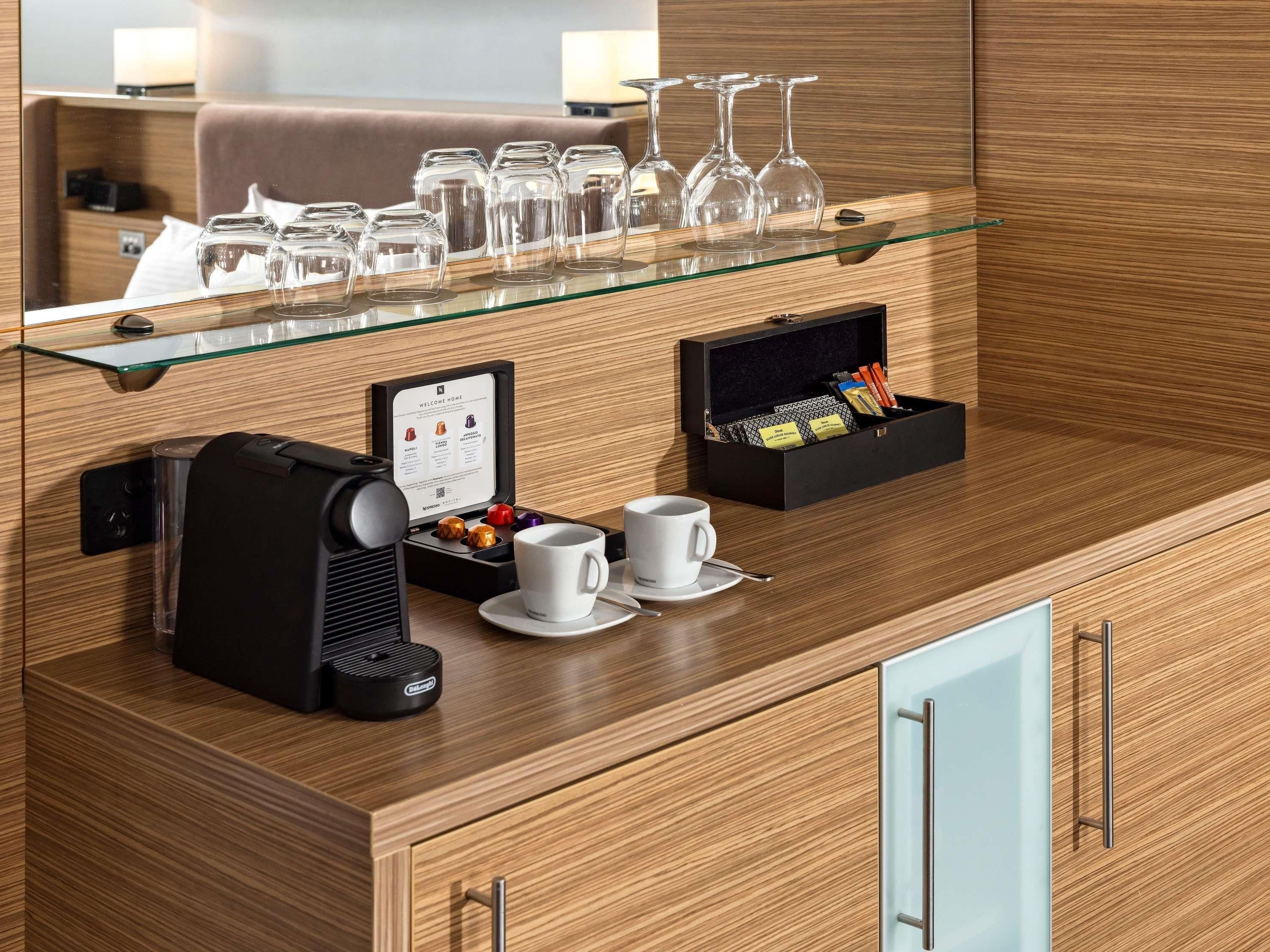 Zona de recepción de un hotel con una máquina de café, una vitrina con copas de vino, una taza de café y una caja de bombones.