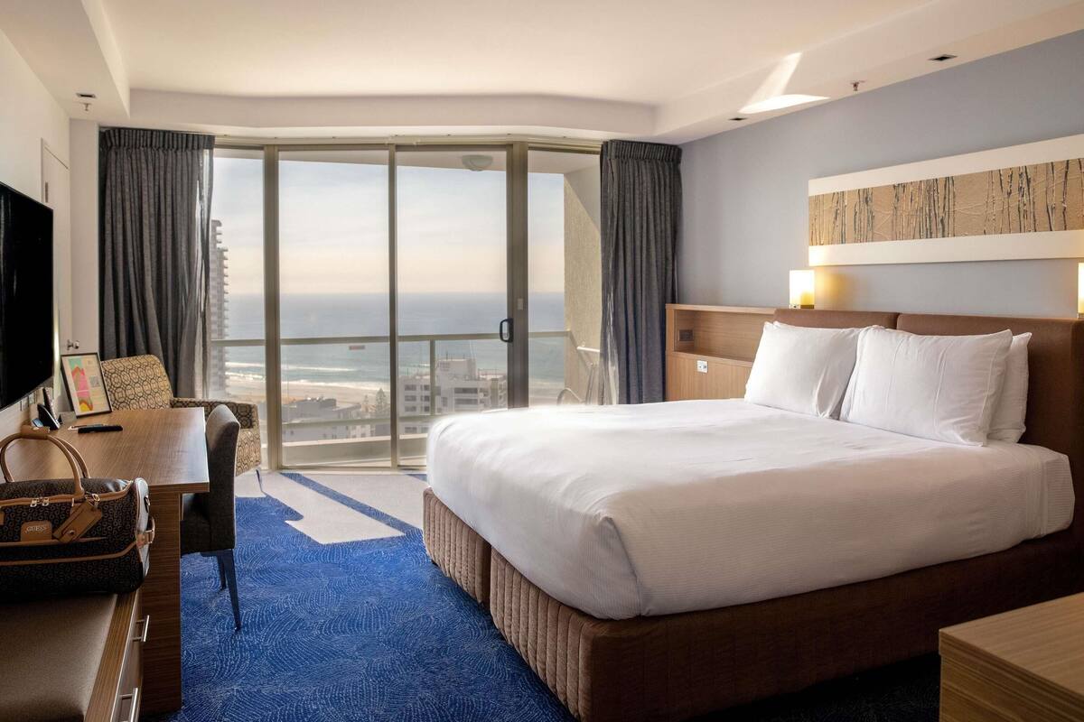 A hotel room with a large bed, a desk, a chair, a TV, and a view of the ocean.