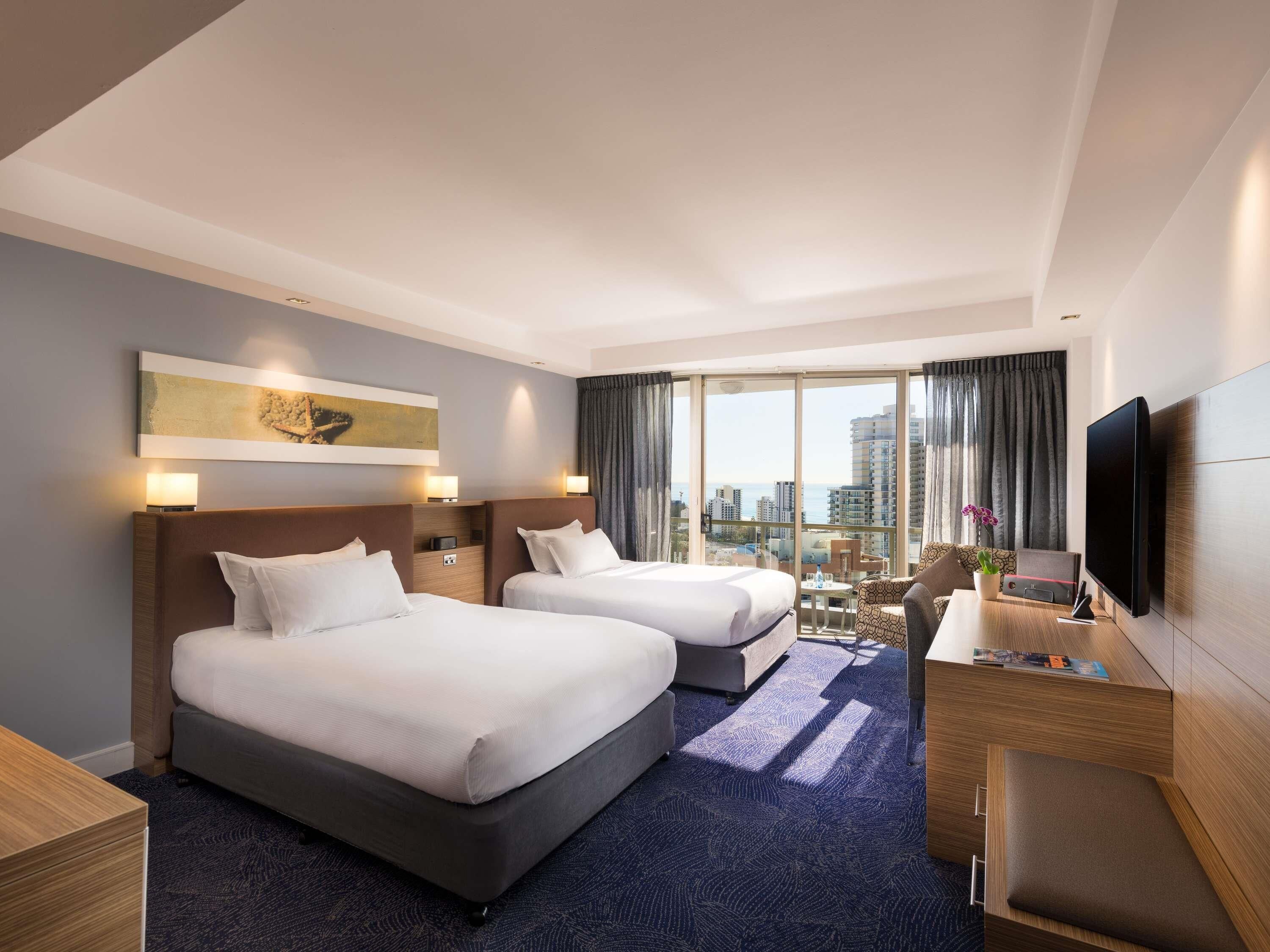 A hotel room with two beds, a desk with a flat-screen TV, a chair, and a large window with a city view.