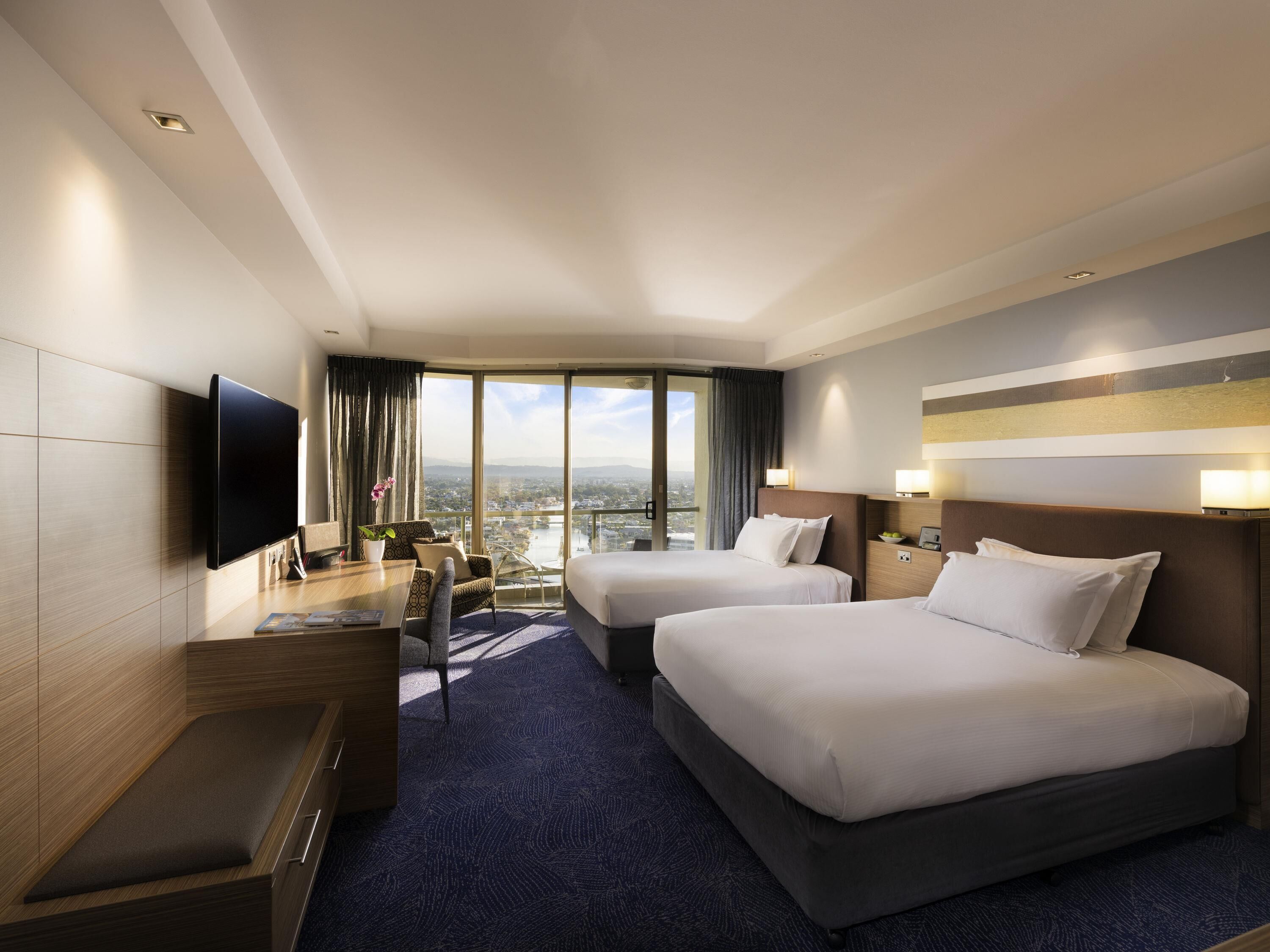 A hotel room with two beds, a flat-screen TV, a desk with a chair, a view of the city, and a large window.