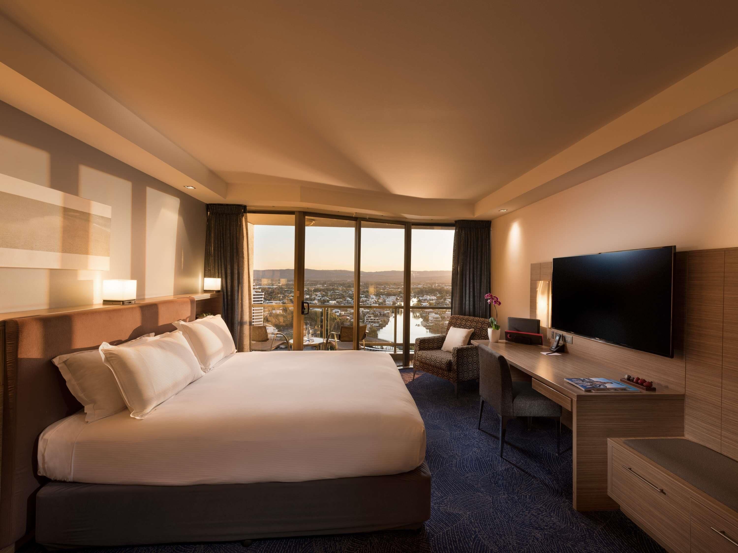 A hotel room with a large bed, a desk, a flat-screen TV, and a view of the city.