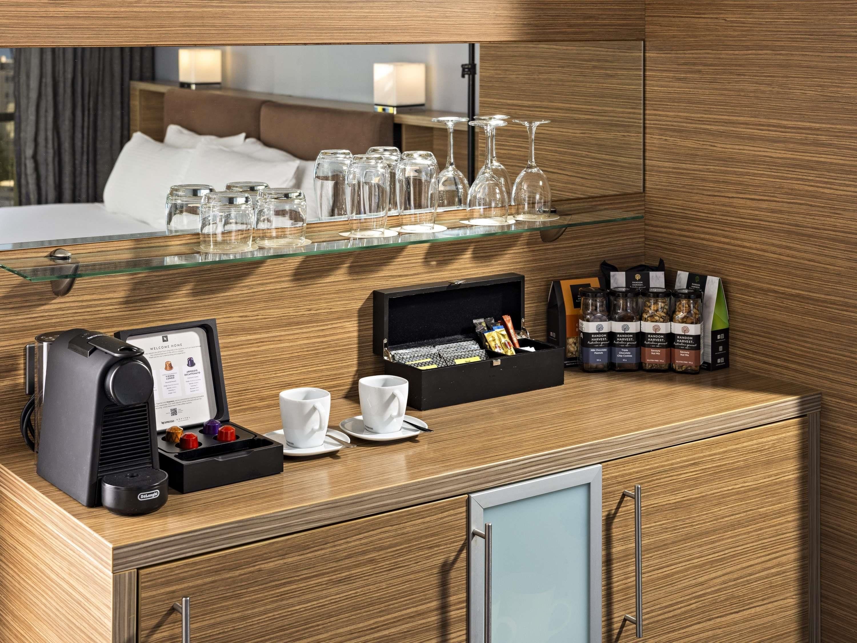 Habitación de hotel con minibar, cafetera, estante de vidrio con copas de vino y una variedad de aperitivos y condimentos.