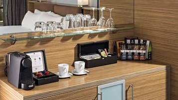 Habitación de hotel con minibar, cafetera, estante de vidrio con copas de vino y una variedad de aperitivos y condimentos.