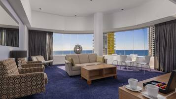 Une chambre d’hôtel moderne dotée d’une grande fenêtre offrant une vue sur la mer et un immeuble de grande hauteur. On y trouve un ensemble de canapés, une table basse et un bureau avec un ordinateur portable et une assiette de pâtisseries.