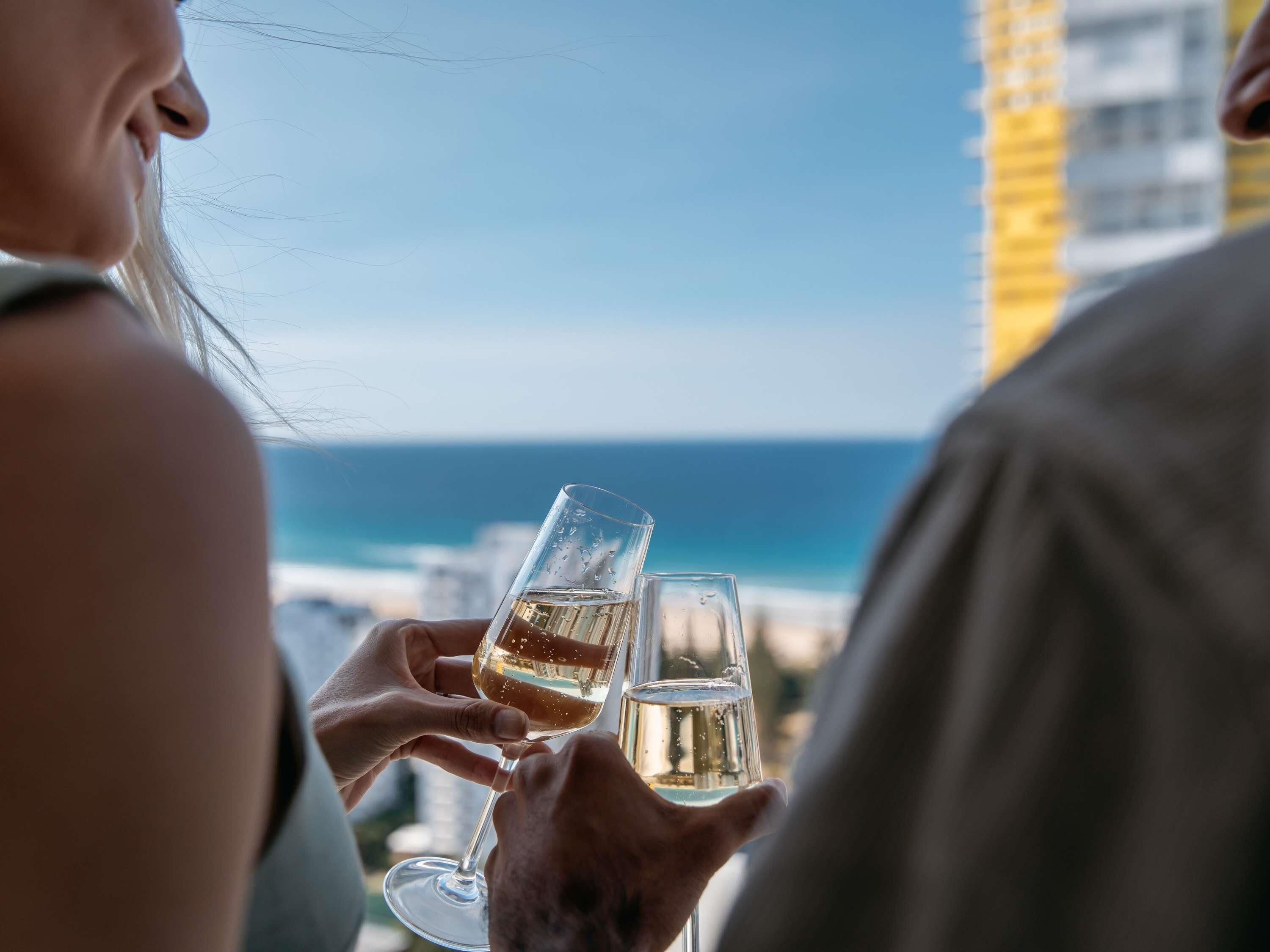 Two people holding glasses of white wine, overlooking a coastal view.
