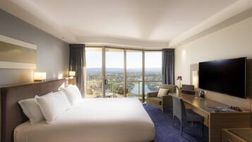 Una habitación de hotel con una cama grande, un escritorio con televisor de pantalla plana, vistas a la ciudad a través de amplios ventanales y un balcón con mesa y sillas.