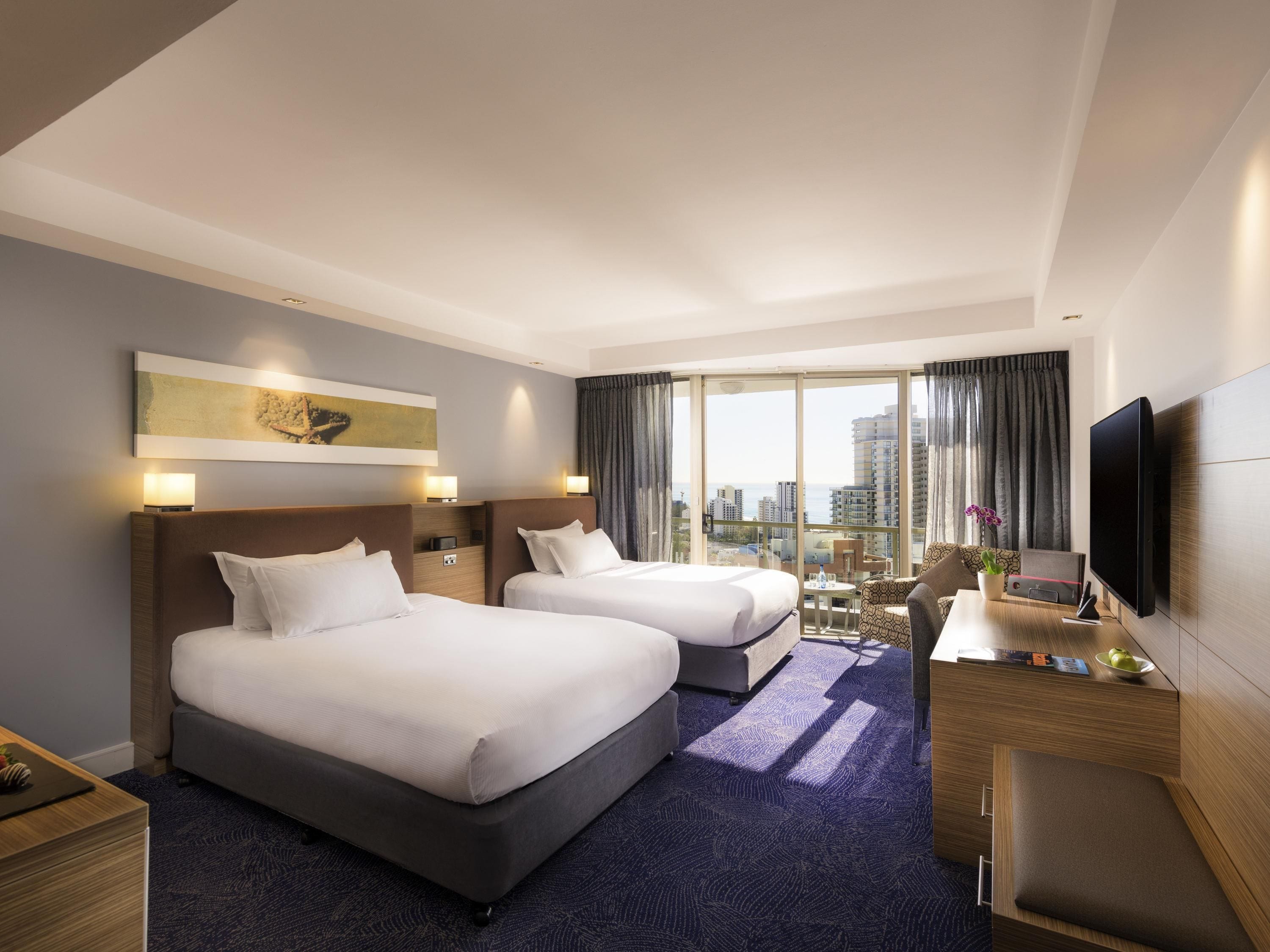 A hotel room with two beds, a desk, a TV, and a city view.