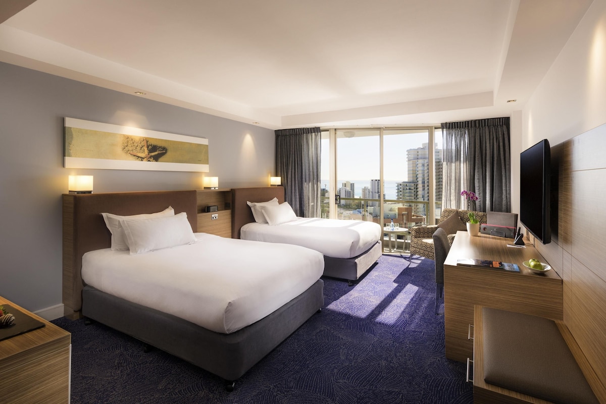 A hotel room with two beds, a desk, a TV, and a city view.