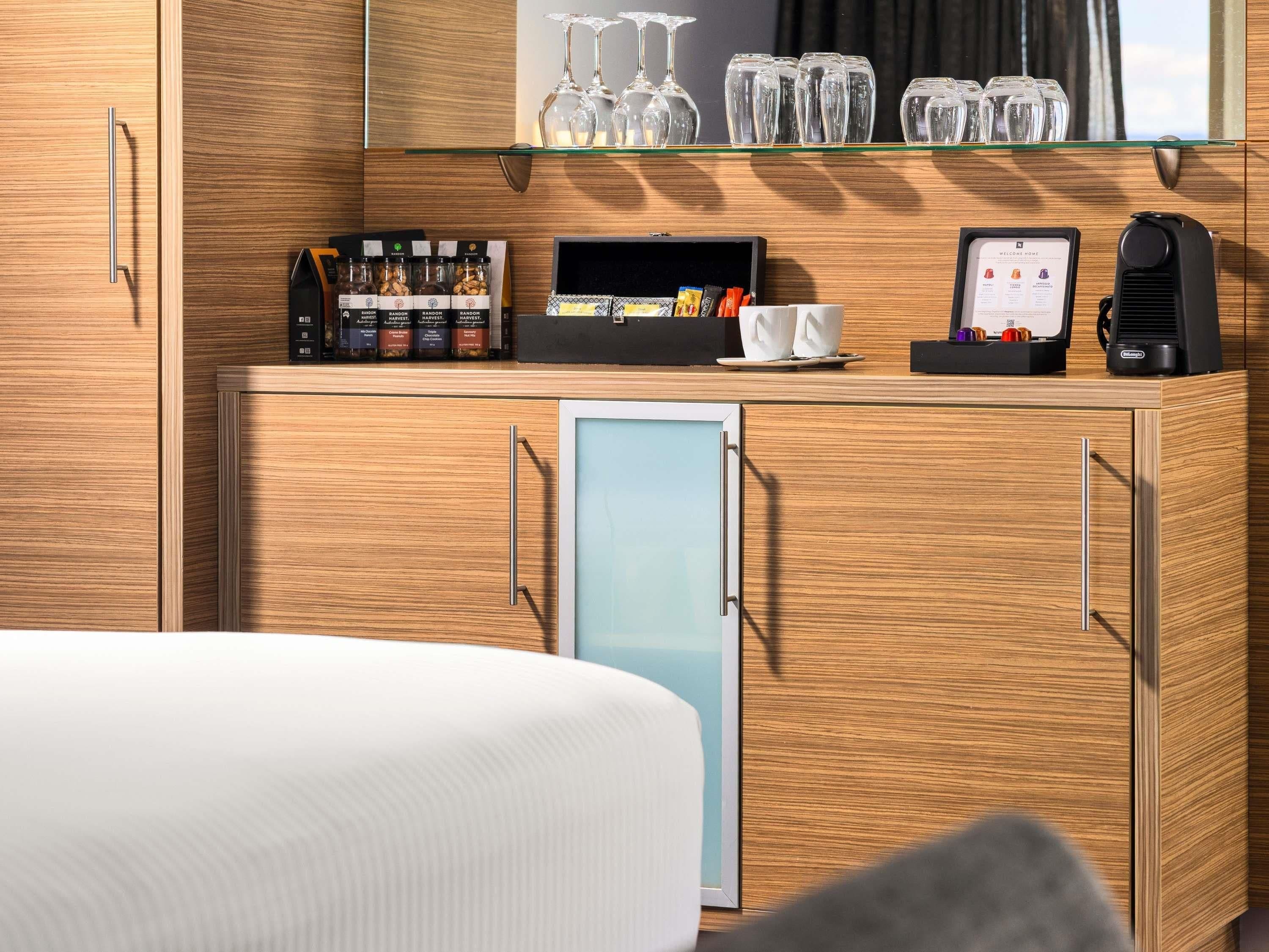 A hotel room with a bed, a minibar, a coffee machine, and a glass display case with wine glasses.