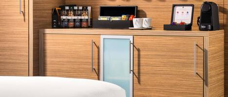 A hotel room with a bed, a minibar, a coffee machine, and a glass display case with wine glasses.