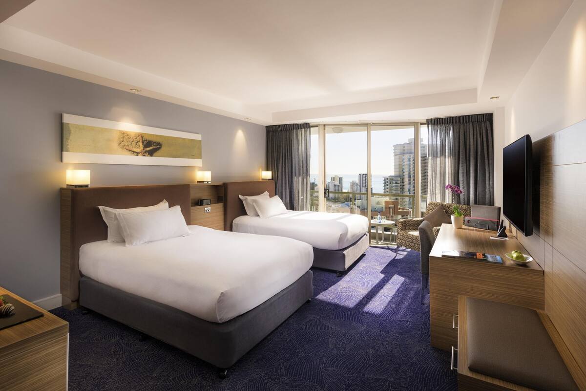 A hotel room with two beds, a desk, a TV, and a city view.