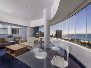 A modern hotel room with a glass table, white chairs, a sofa, and a large window offering a city and ocean view. - Sofitel Gold Coast Broadbeach (Broadbeach)