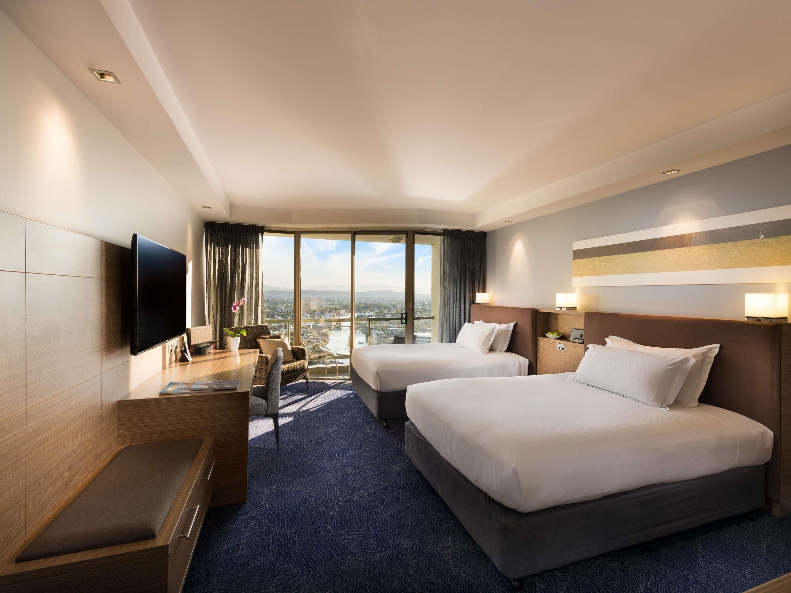 A hotel room with two beds, a desk, a chair, a TV, and a large window offering a city view.