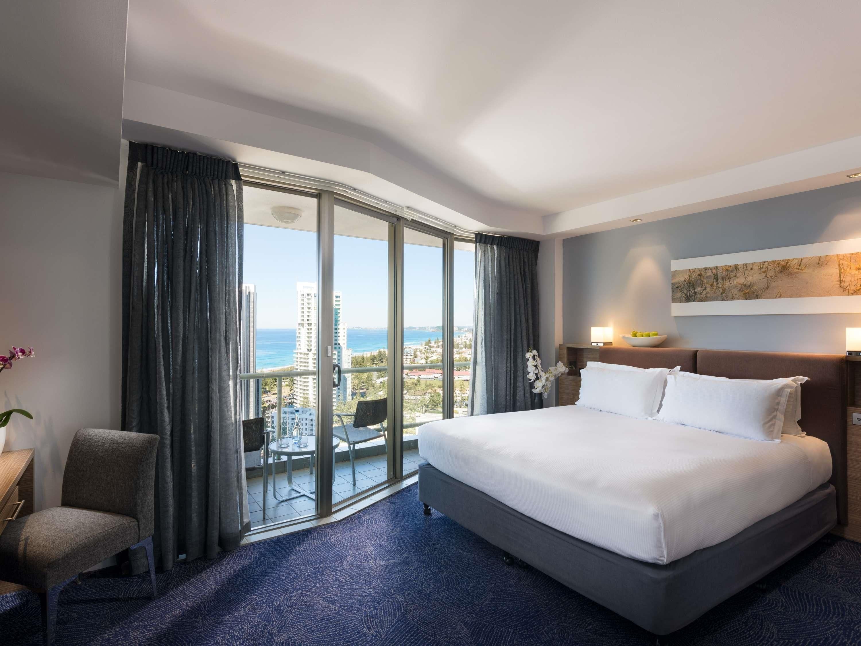 A hotel room with a large bed, a chair, a balcony with a view of the city and sea, and a painting on the wall.