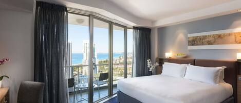A hotel room with a large bed, a chair, a balcony with a view of the city and sea, and a painting on the wall.
