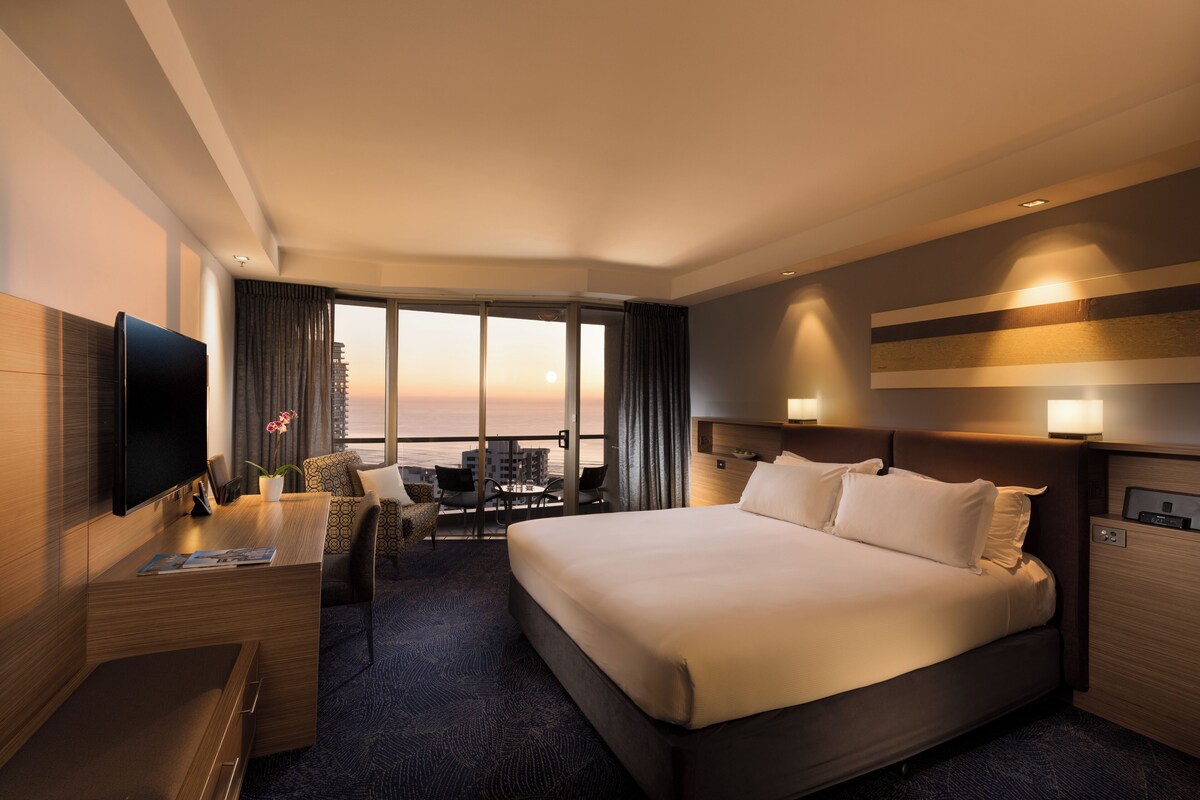 Une chambre d’hôtel moderne dotée d’un grand lit, d’un bureau avec un téléviseur à écran plat, d’un coin salon offrant une vue sur le coucher de soleil et d’un balcon.