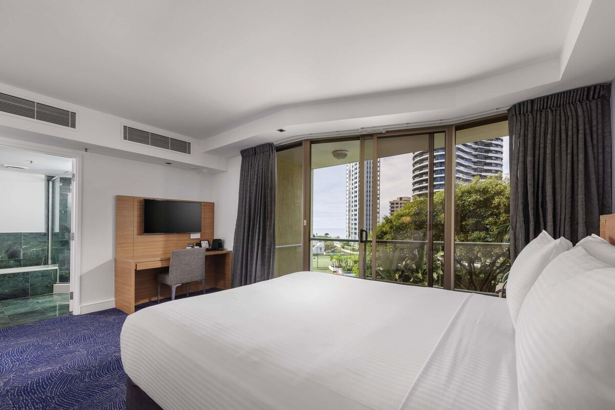 A hotel room with a large bed, a desk with a chair, a TV, and a balcony with a view of buildings and trees.