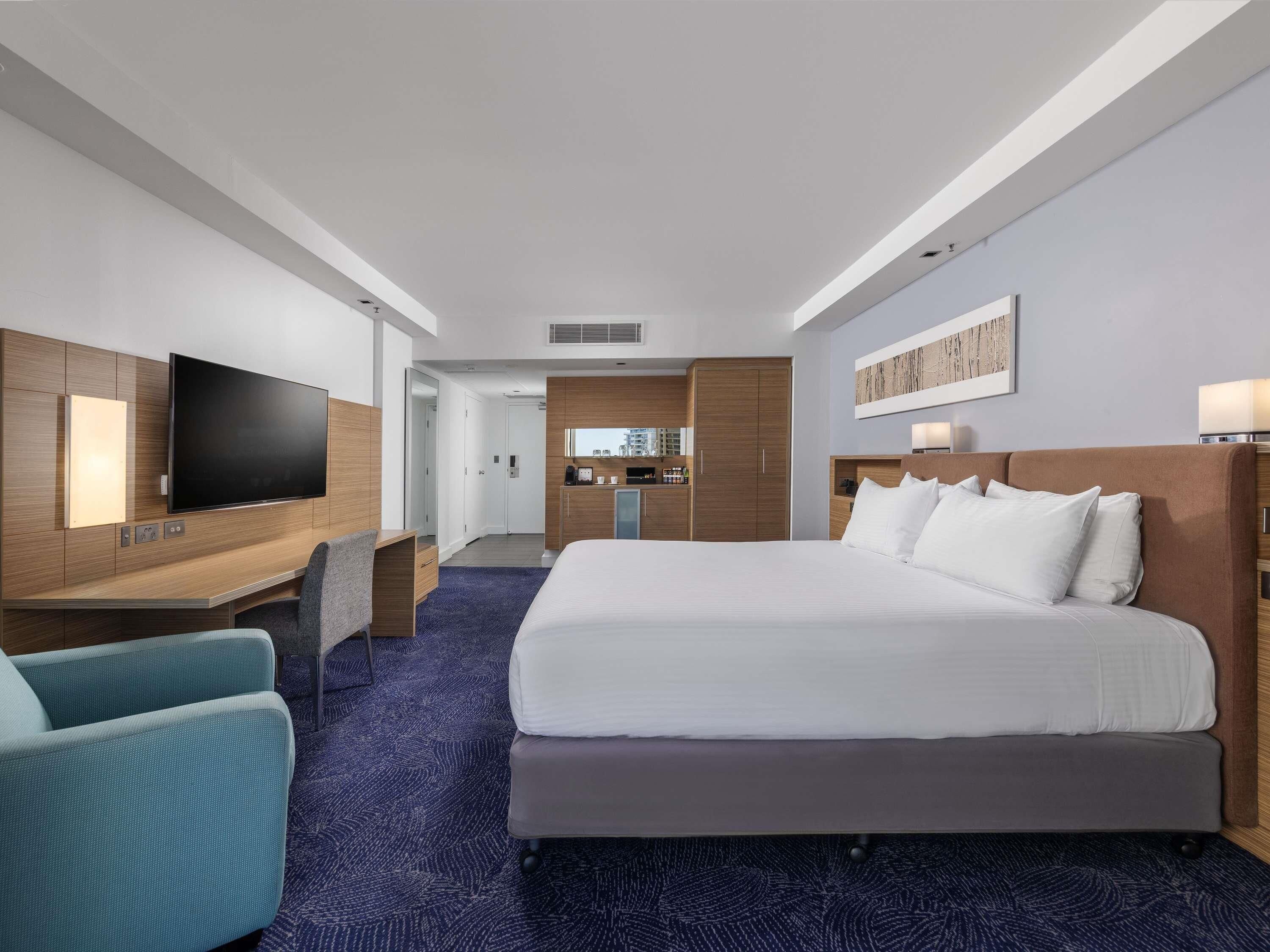 A hotel room with a large bed, a desk with a flat-screen TV, a seating area with a chair, and a kitchenette in the background.