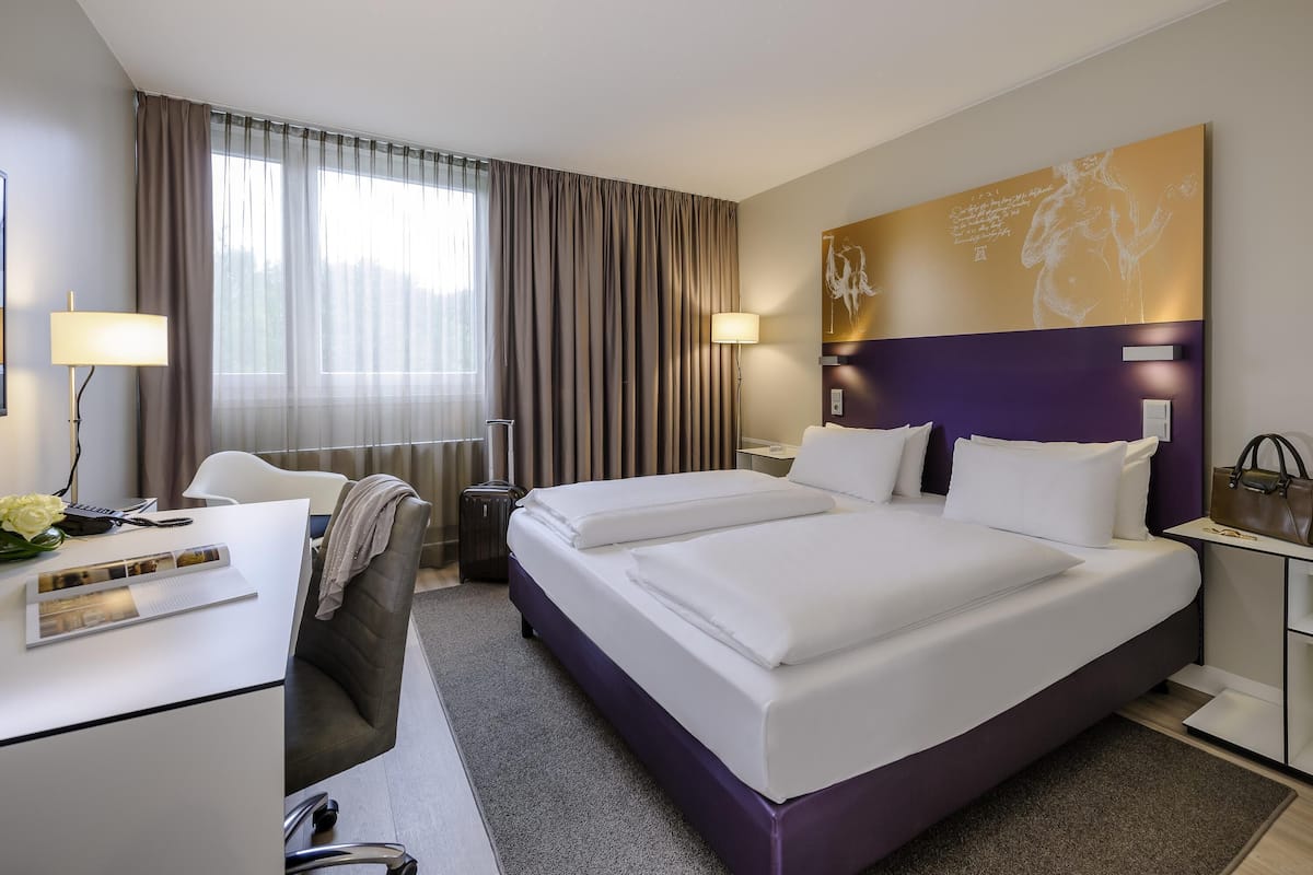 A hotel room with a large bed, a desk with a chair, a lamp, and a window with curtains.