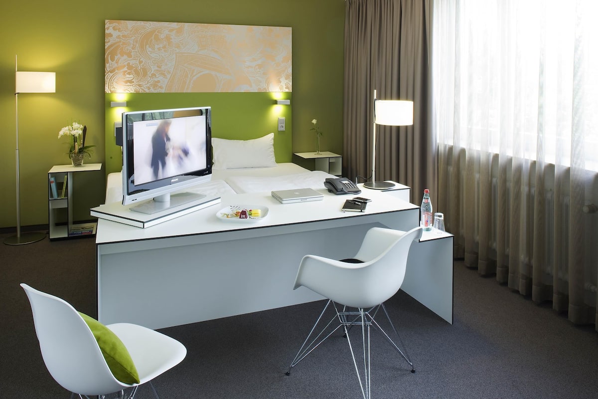 Habitación de hotel con cama, escritorio con computadora, silla, televisor y ventana con cortinas.